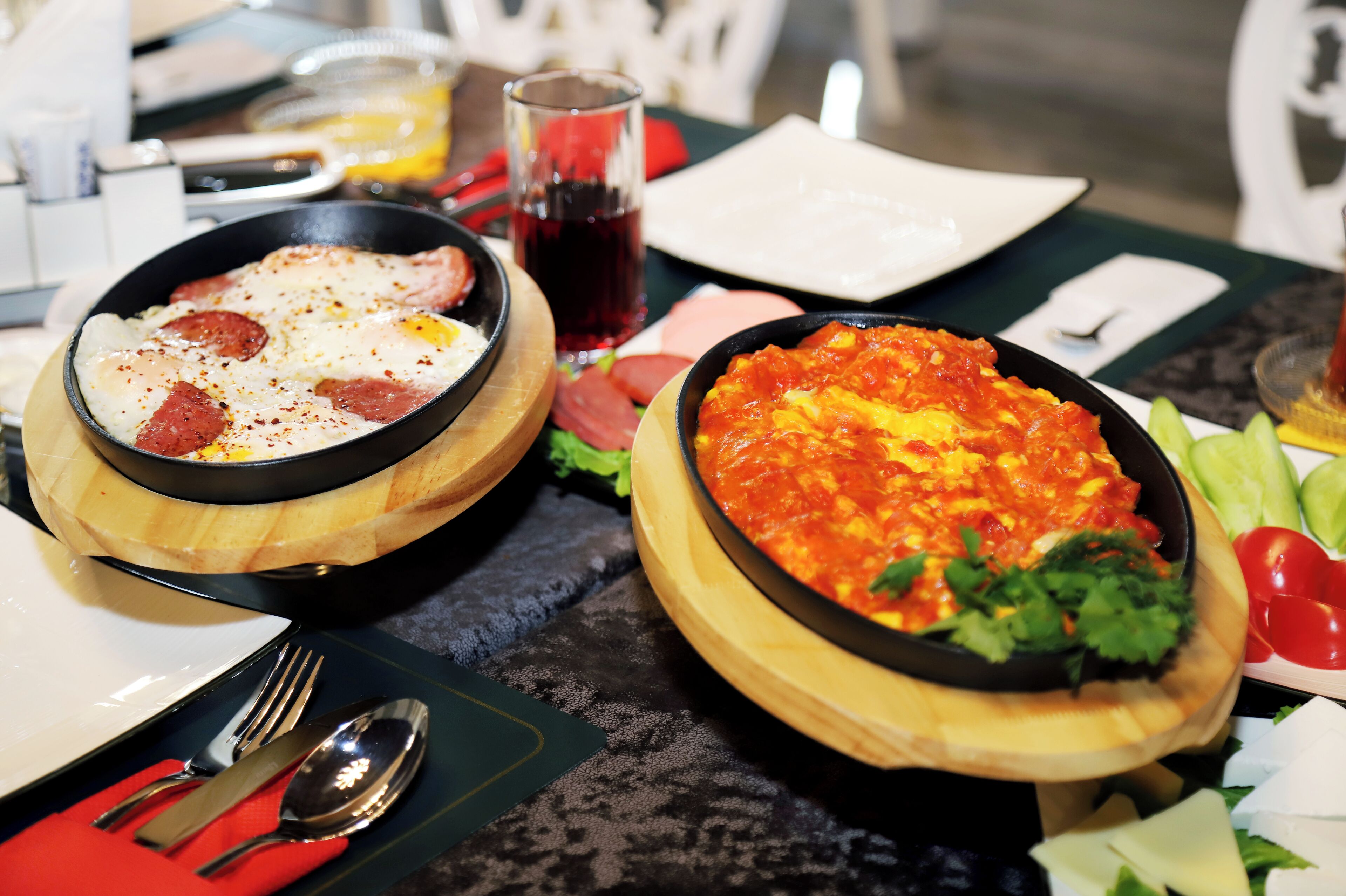 Una mesa de comedor con dos platos de comida, un vaso de bebida roja, y un plato de tomates y pepinos rebanados.