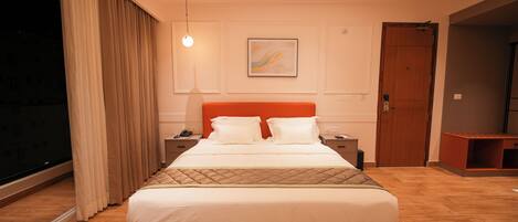 A hotel room with a large bed, a bedside table, a nightstand, a window with curtains, a door, and a ceiling light.