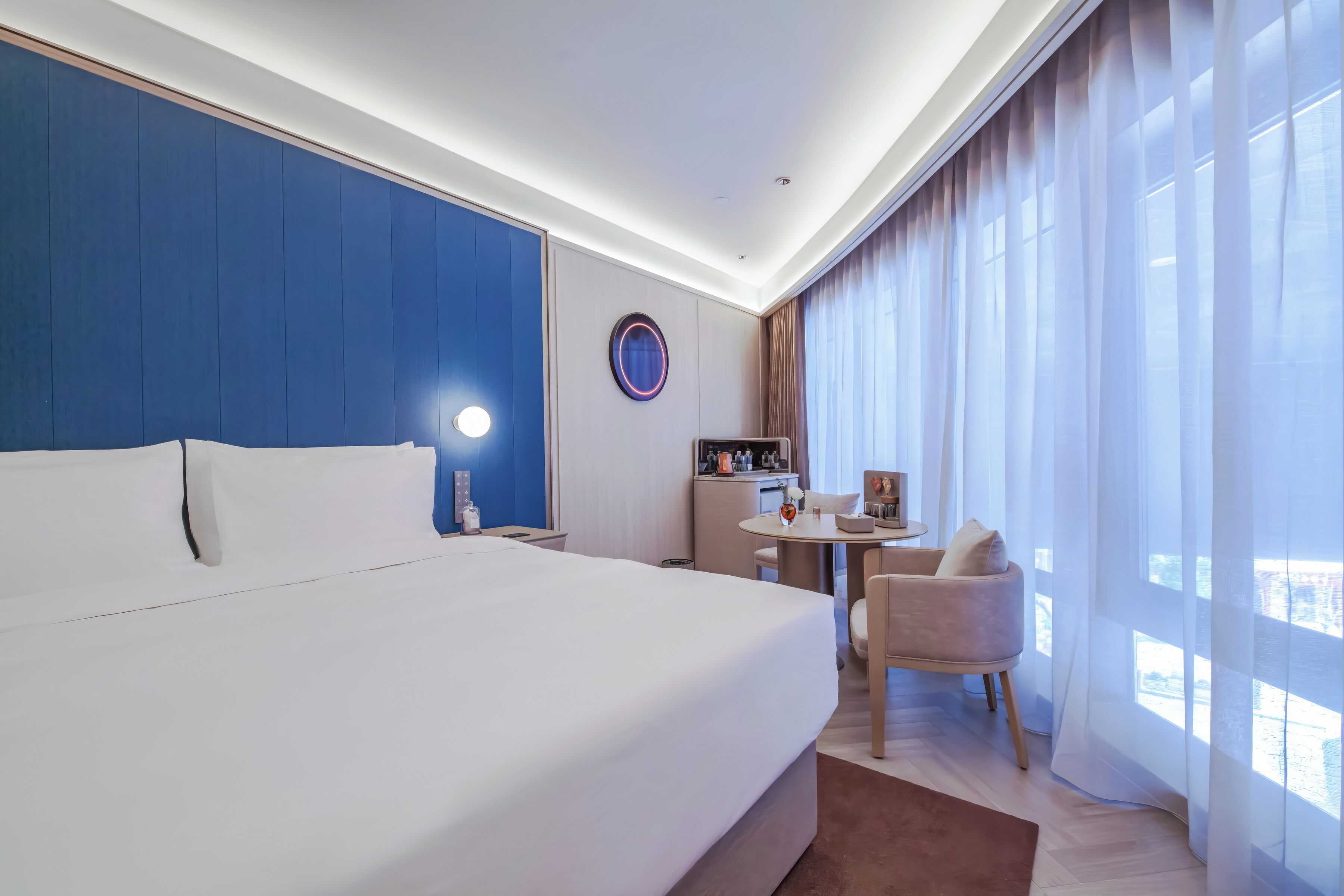 A hotel room with a large bed, a desk, a chair, a round mirror, and a view of the city through the window.