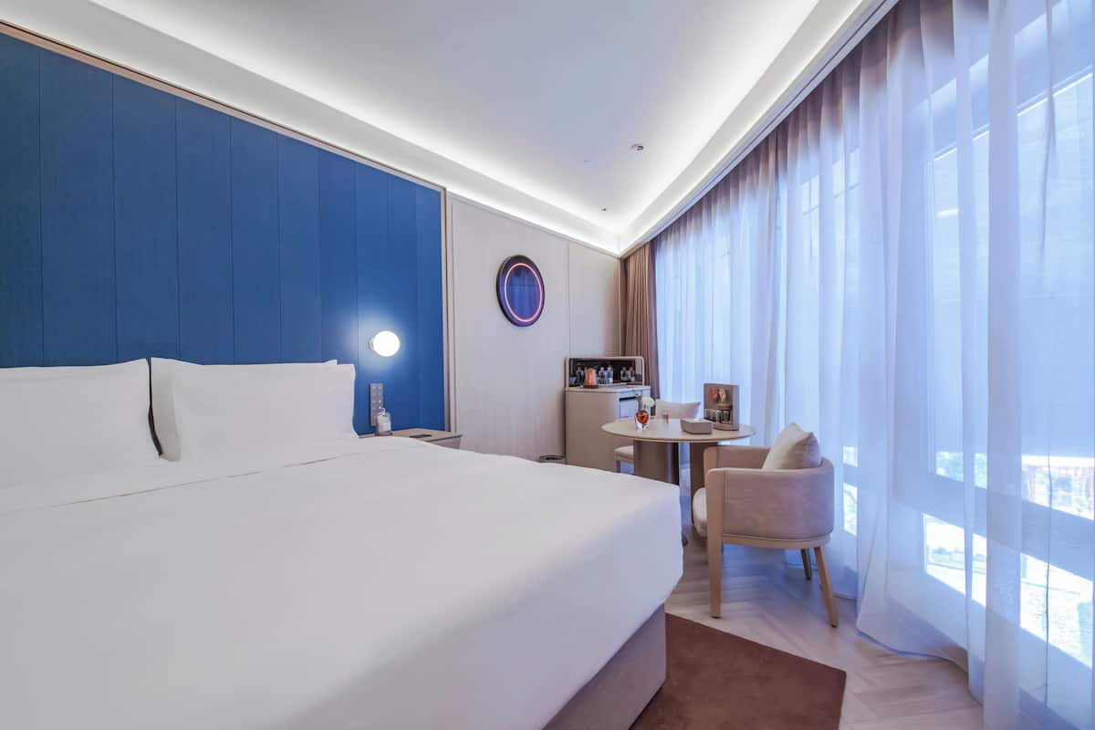 Habitación de hotel con una cama grande, un escritorio, una silla, un espejo redondo y vistas a la ciudad a través de la ventana.