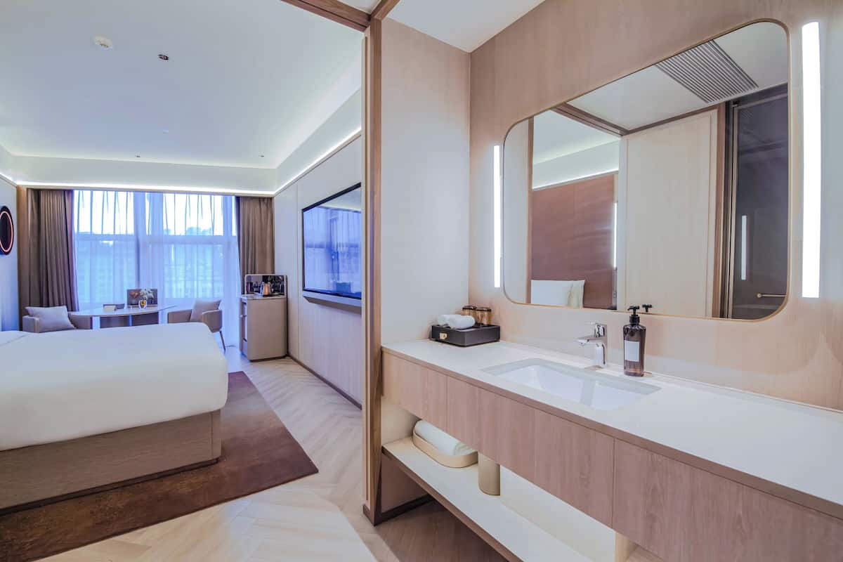 Habitación de hotel moderna con una cama grande, baño con espejo y lavamanos, escritorio y vistas a la ciudad.