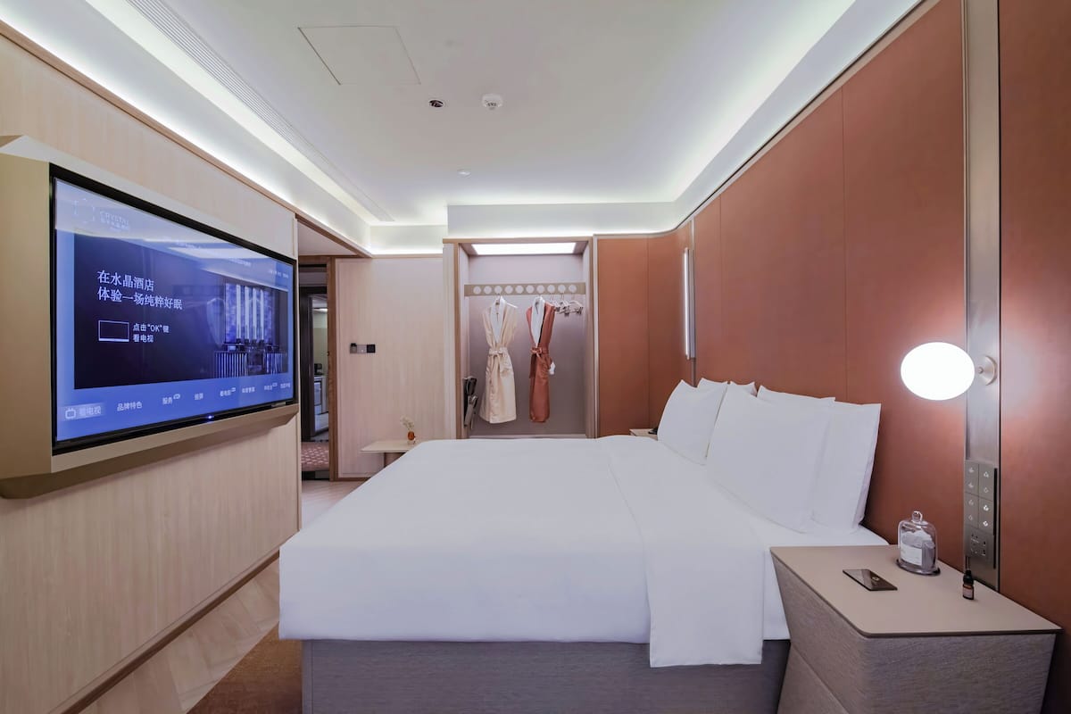 Habitación de hotel con un televisor de pantalla plana grande montado en la pared, una cama con ropa blanca, una mesita de noche con una lámpara y un espejo.