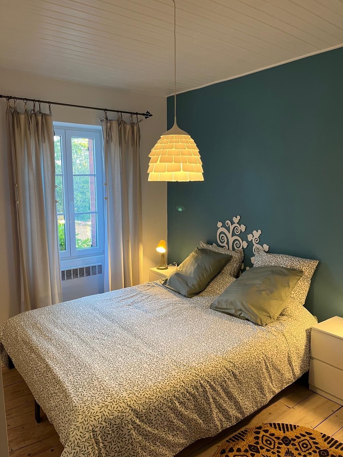 A bedroom with a bed, curtains, a hanging lamp, a bedside table, and a window.
