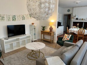 Un salon comprenant un téléviseur, une table ronde en marbre, un canapé, un coin repas avec une table et des chaises, une cuisine à l’arrière-plan et un luminaire décoratif au plafond.
