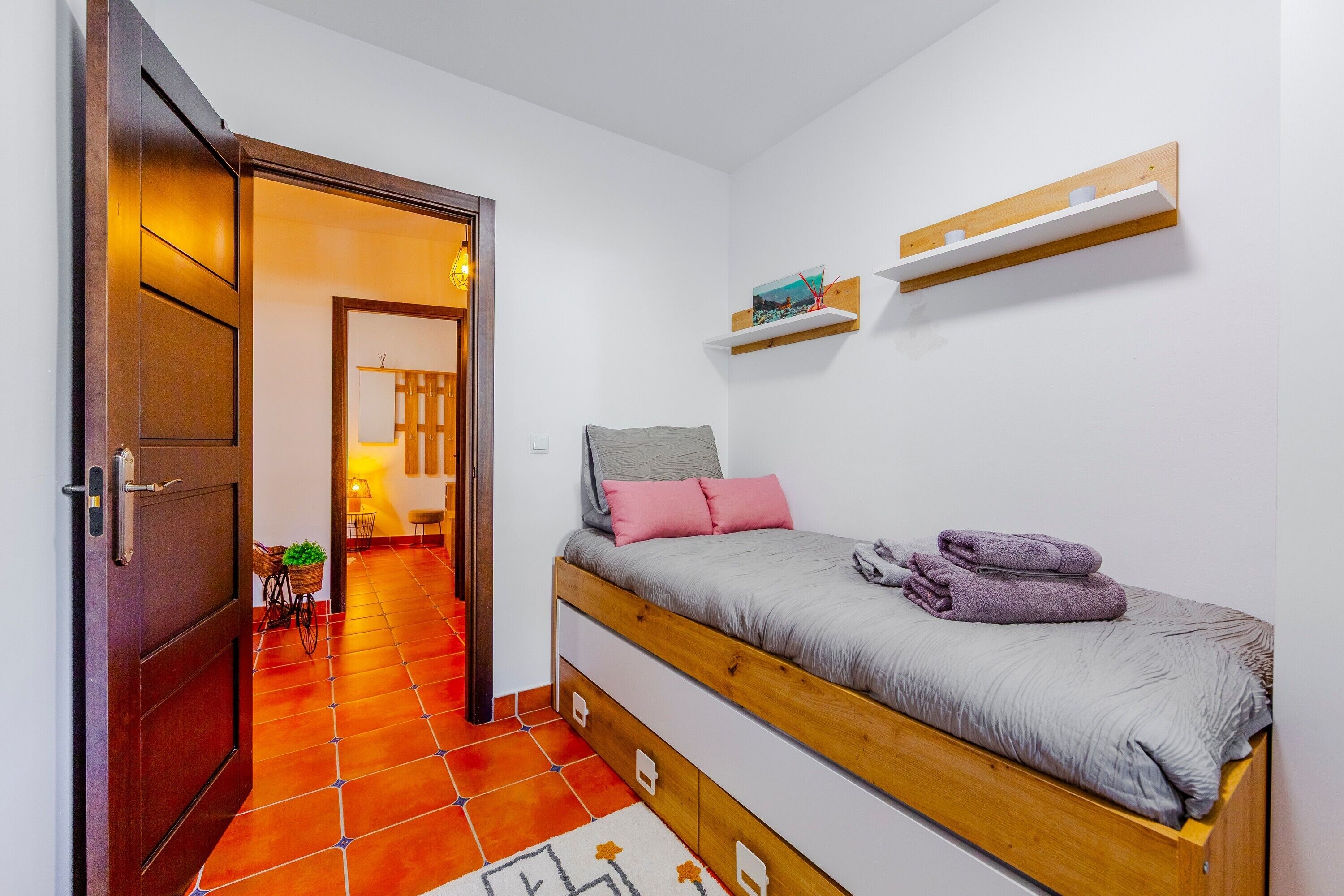 Un dormitorio con cama, una puerta de madera, un pasillo y un estante fijado a la pared.