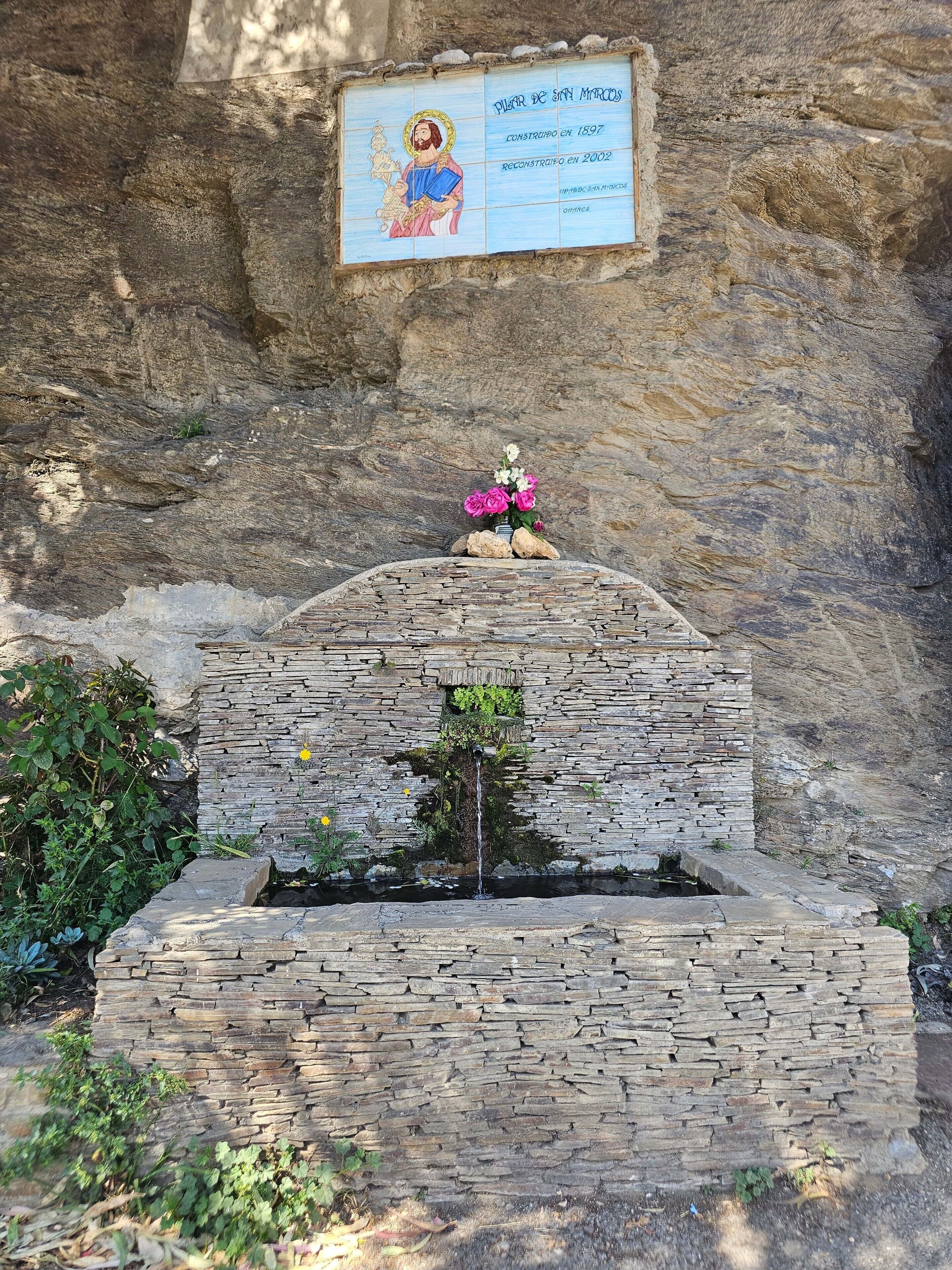 Uma construção de pedra com um ícone religioso, uma placa e uma pequena fonte.