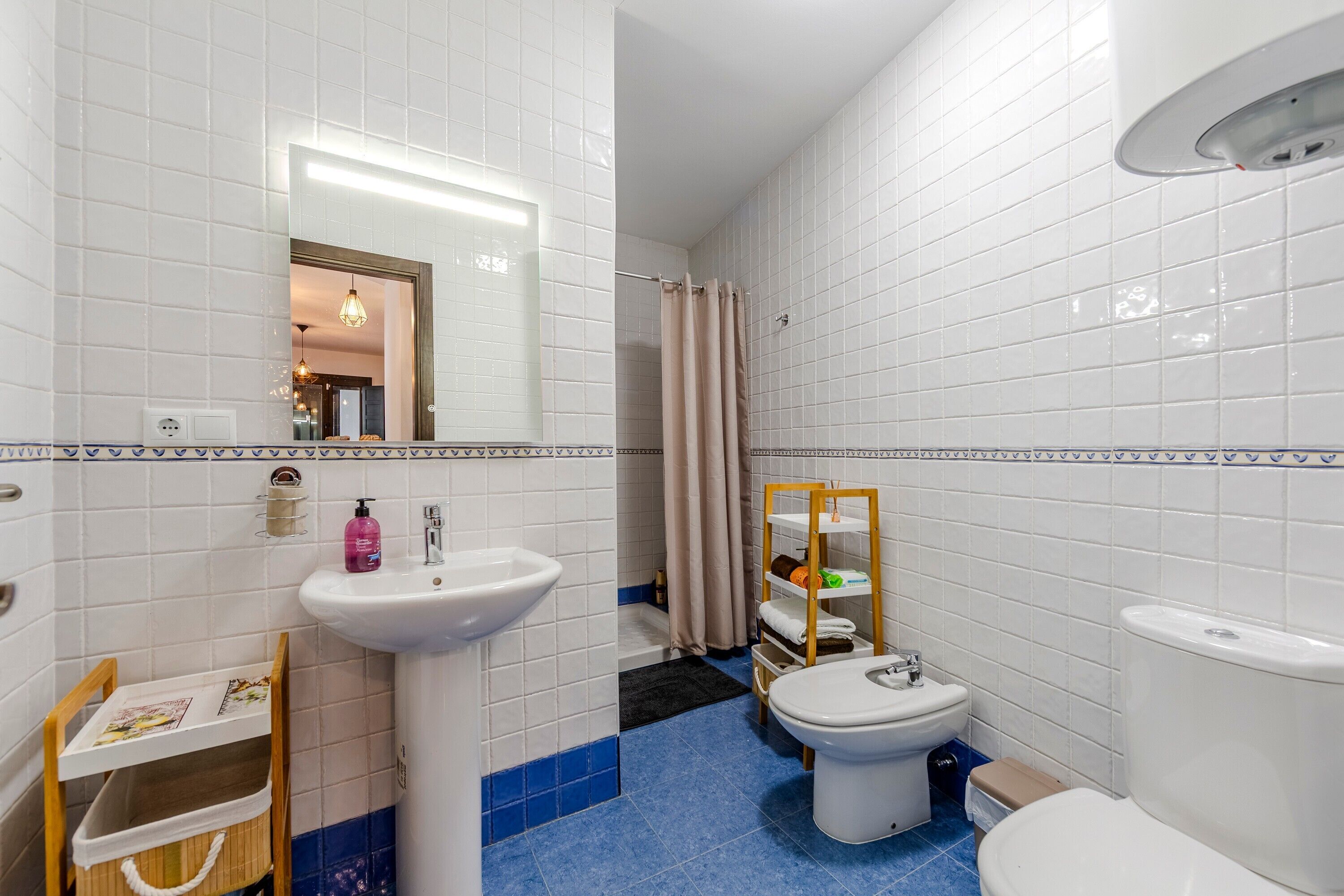 Un baño con paredes de azulejos blancos, un lavamanos, un inodoro y un bidé. Hay un espejo, un estante con artículos de aseo y una silla de madera.