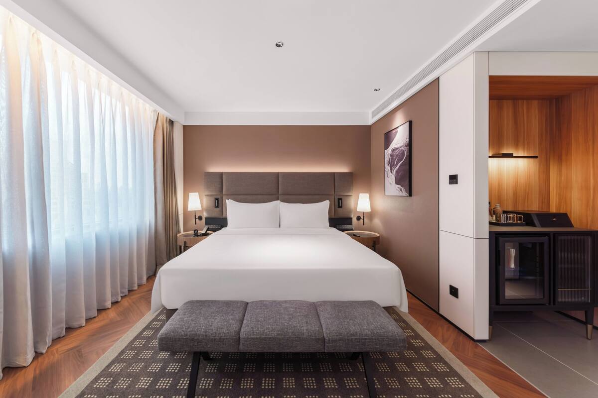 A hotel room with a large bed, bedside lamps, a bench, a desk, and a view of the outside.