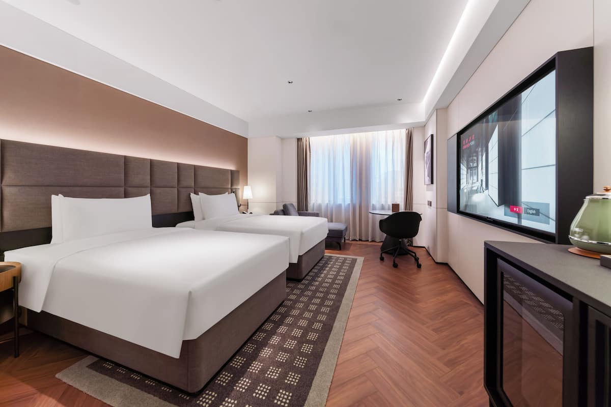 A hotel room with two beds, a desk with a TV, a chair, and a window with curtains.