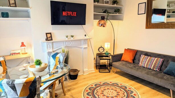 A living room with a sofa, a TV mounted on the wall, a fireplace, a dining table with chairs, and a patterned rug. - Harold House (Leeds)