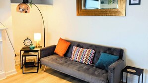 A grey sofa with colorful cushions, a small side table, a round patterned rug, and a framed picture on the wall. - Harold House (Leeds)