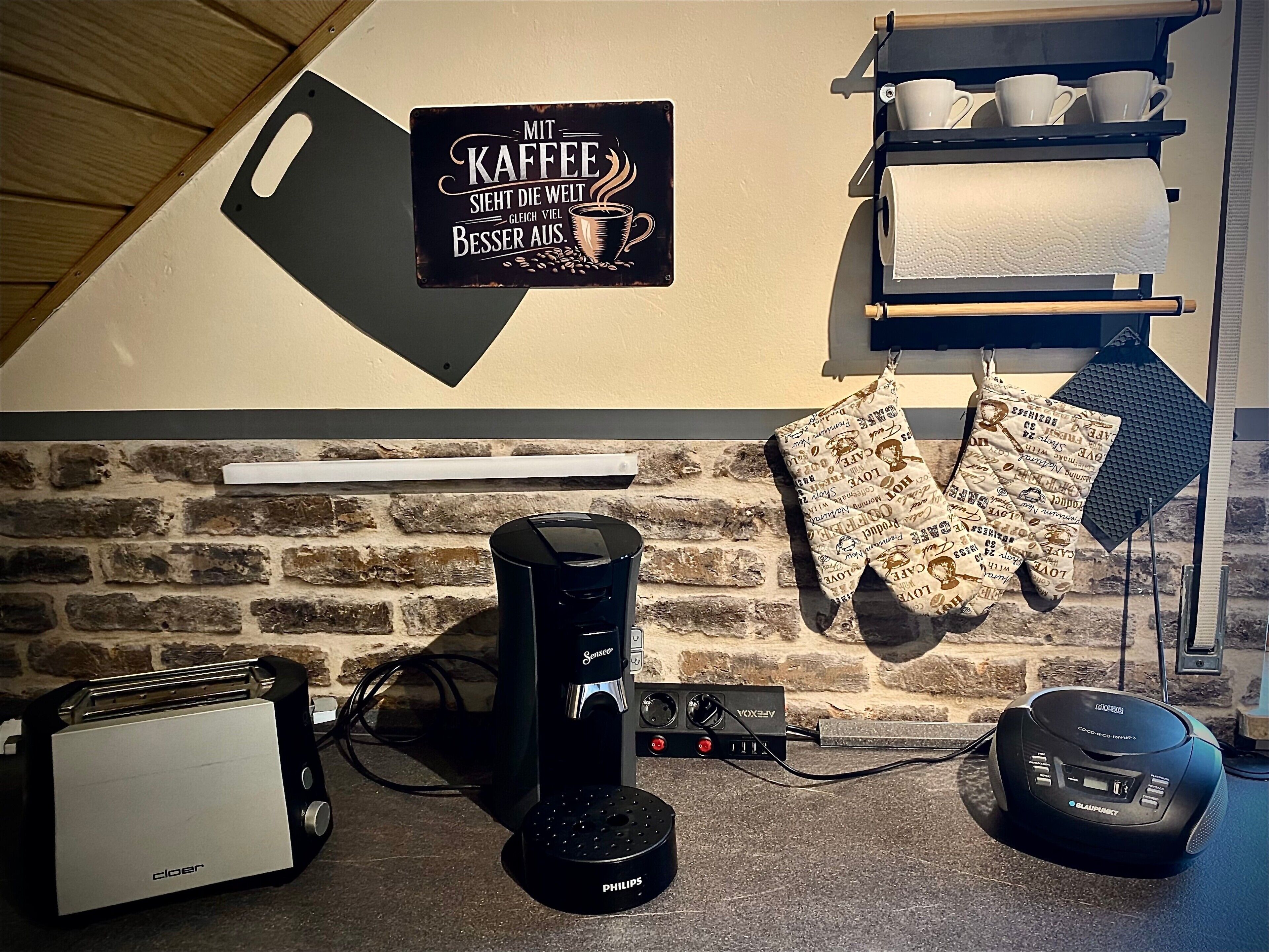 A kitchen with a coffee machine, toaster, and kettle, a brick wall, and a sign that says 'Mit Kaffee sieht die Welt besser aus.'