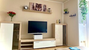A modern living room with a flat-screen TV, wooden cabinets, a vase of flowers, and a plant on a shelf. - Apartment 'Bassd Scho' with Shared Garden, Balcony and Wi-Fi (Ebensfeld)