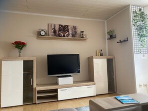 Ein Wohnzimmer mit Flachbildfernseher, Holzschränken, einer Vase mit roten Blumen und einem Bücherregal mit Dekoration.