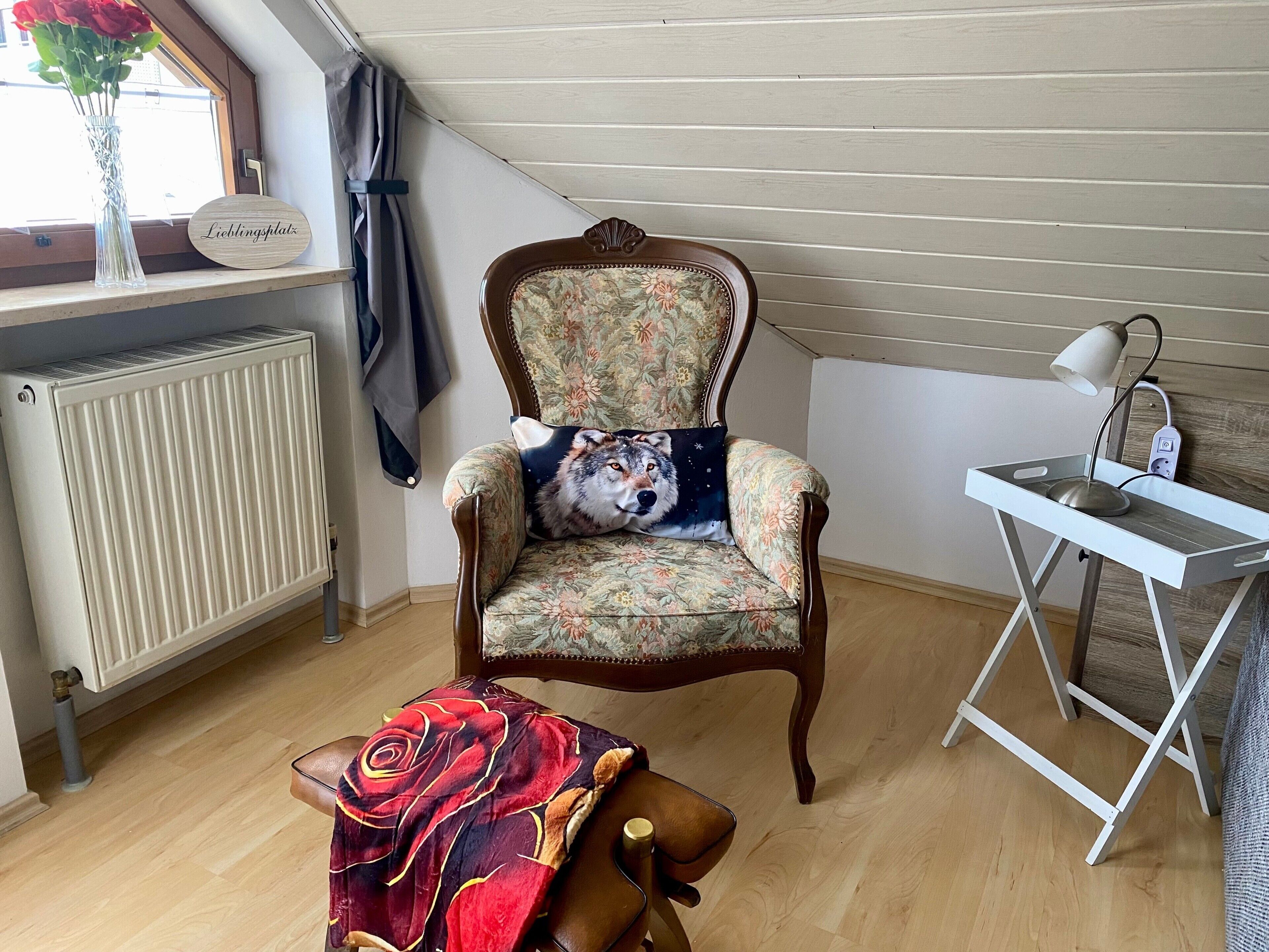 Ein gemütliches Zimmer mit einem blumigen Sessel, einem kleinen Tisch mit Lampe, einem Heizkörper und einem Fenster mit Vorhang.