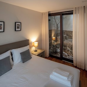 Habitación de hotel con cama, dos almohadas, mesita de noche con lámpara y ventana con cortinas.