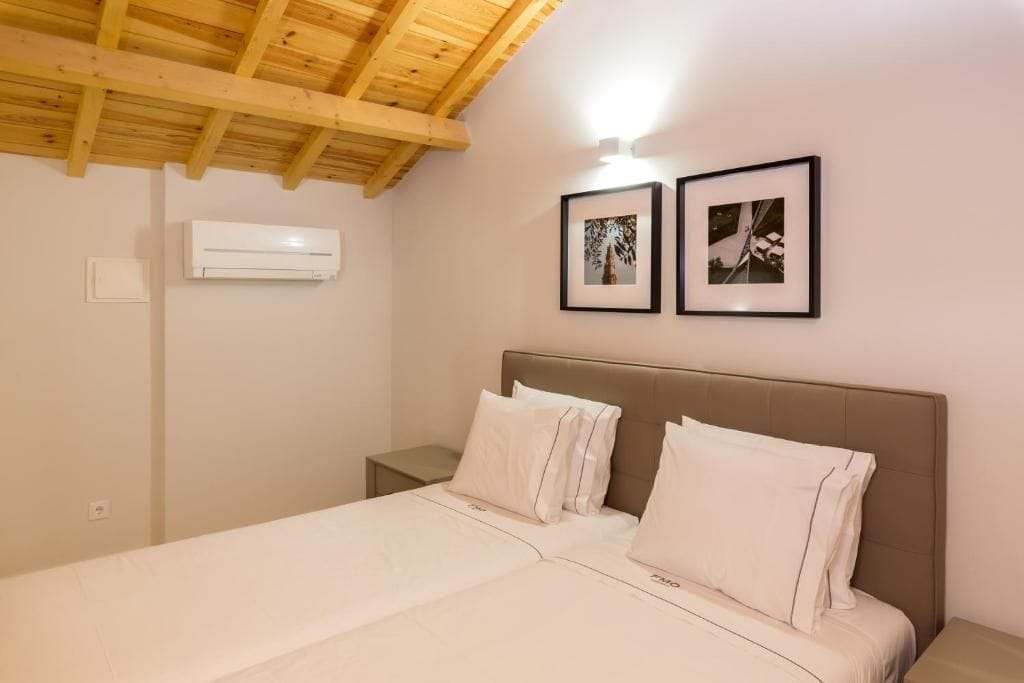 Un dormitorio con techo de madera, una cama con ropa blanca, dos cuadros en la pared y un aire acondicionado.