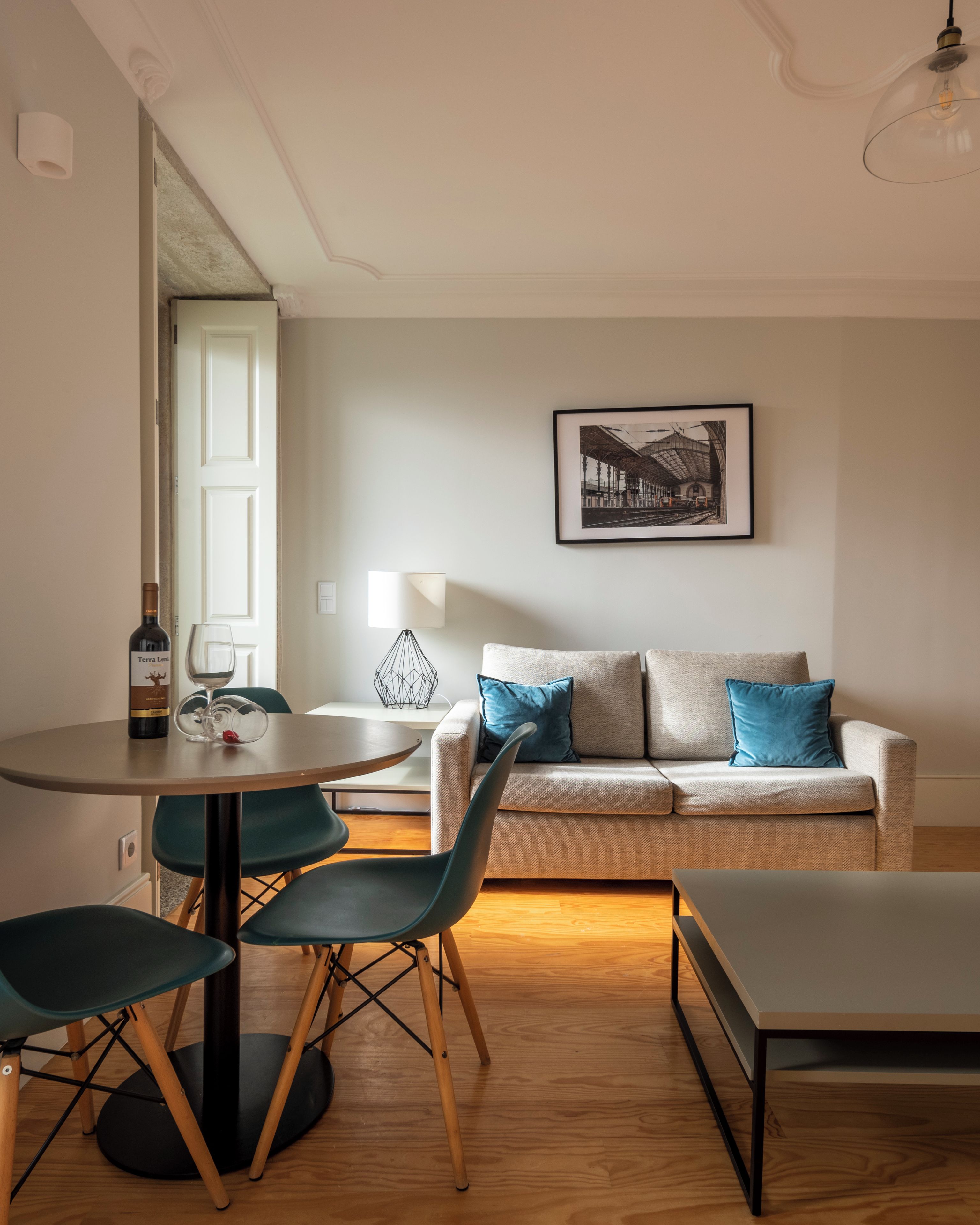 Una sala moderna con una mesa redonda de comedor, dos sillas, un sofá con cojines azules, una lámpara de pie y un cuadro en la pared.