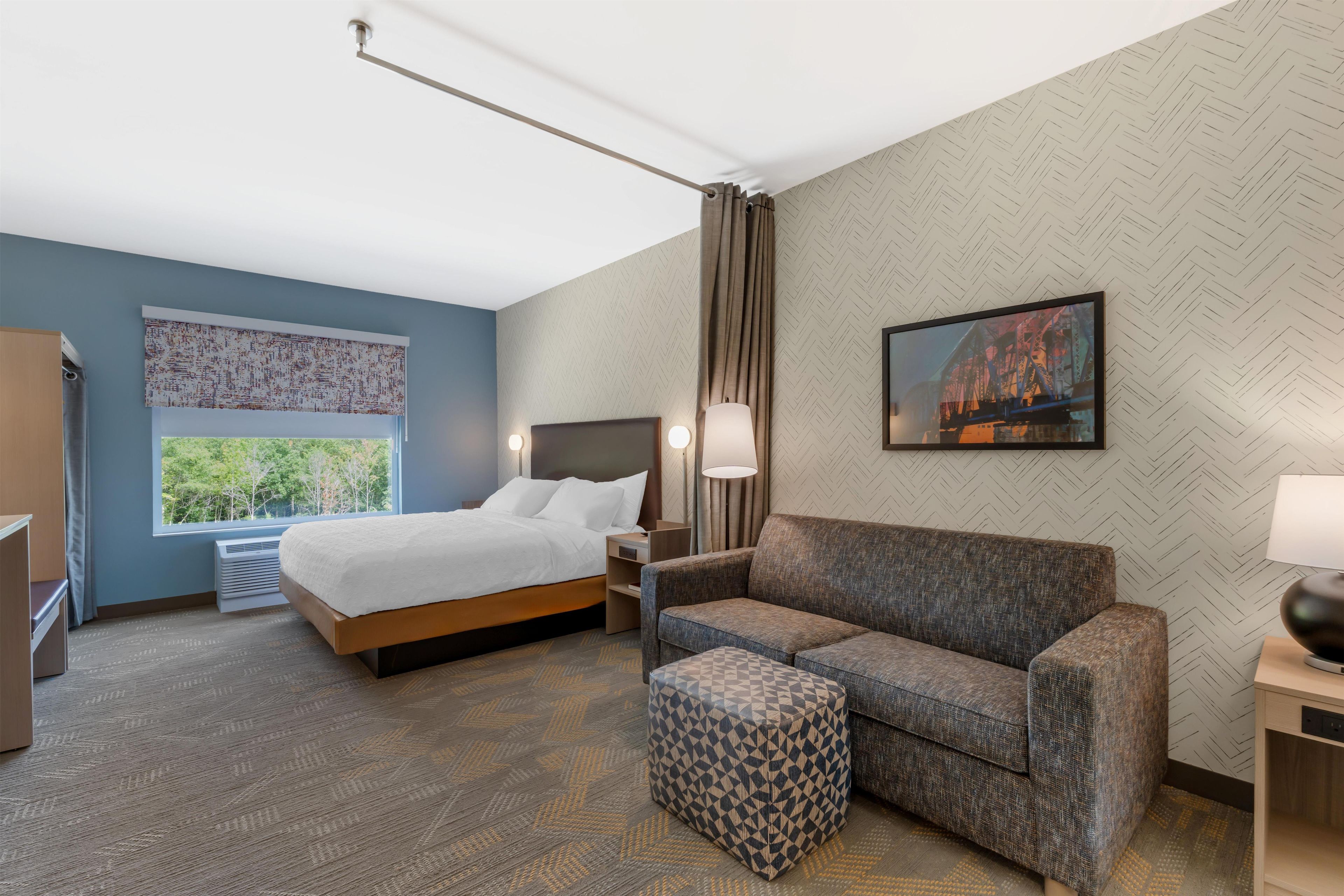 A hotel room with a bed, a sofa, a small ottoman, a nightstand, a lamp, and a window with a view of greenery.