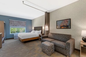 Habitación de hotel con cama, sofá, pequeño taburete, mesita de noche, lámpara y ventana con vistas a zonas verdes.