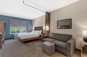 Habitación de hotel con cama, sofá, pequeño taburete, mesita de noche, lámpara y ventana con vistas a zonas verdes.