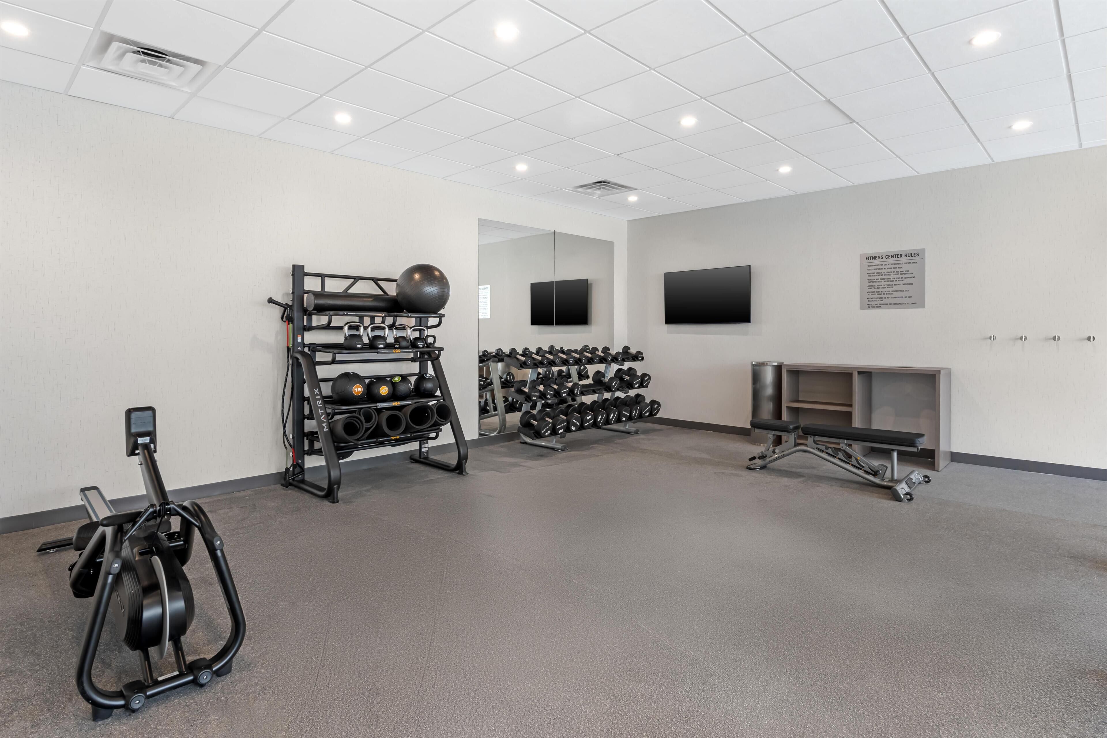 Un gimnasio con equipo de ejercicio, un televisor instalado en la pared y un tablón de anuncios con normas.