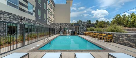 A rectangular outdoor swimming pool with a tiled deck, surrounded by a stone wall and a metal fence. There are lounge chairs and a bar area with yellow chairs.