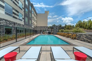 Una piscina rectangular al aire libre con solárium empedrado, rodeada por un muro de piedra y una valla metálica. Hay sillones de descanso y una zona de bar con sillas amarillas.