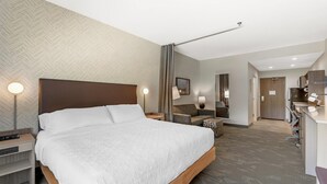 Habitación de hotel con una cama grande, un escritorio, una silla, un espejo y una kitchenette.