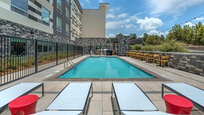 Una piscina rectangular al aire libre con solárium empedrado, rodeada por un muro de piedra y una valla metálica. Hay sillones de descanso y una zona de bar con sillas amarillas.