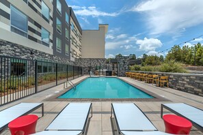 A rectangular outdoor swimming pool with a tiled deck, surrounded by a stone wall and a metal fence. There are lounge chairs and a bar area with yellow chairs.