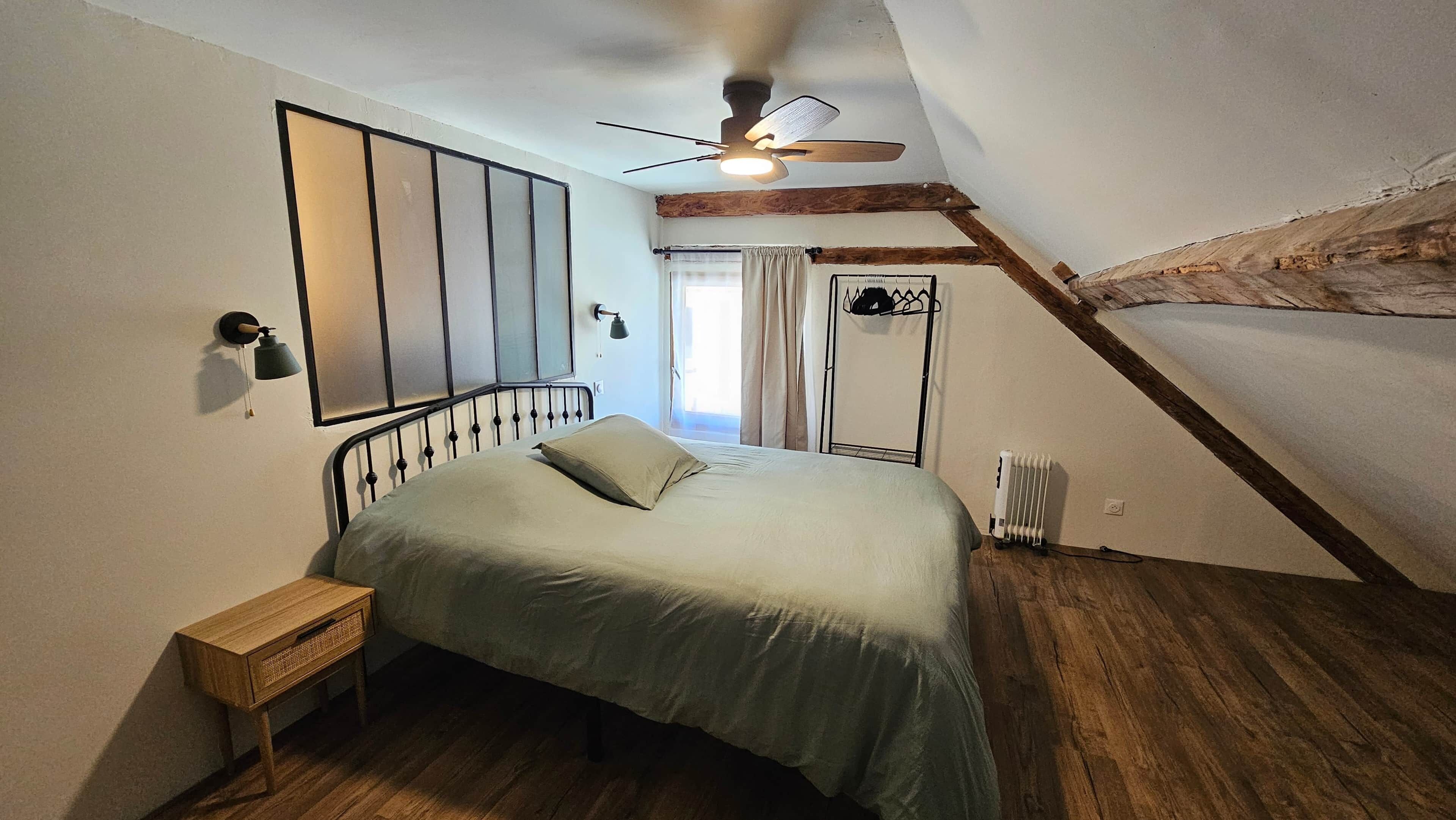 Une chambre à coucher comprenant un lit, une table de chevet, un ventilateur de plafond, une fenêtre avec des rideaux et un radiateur.