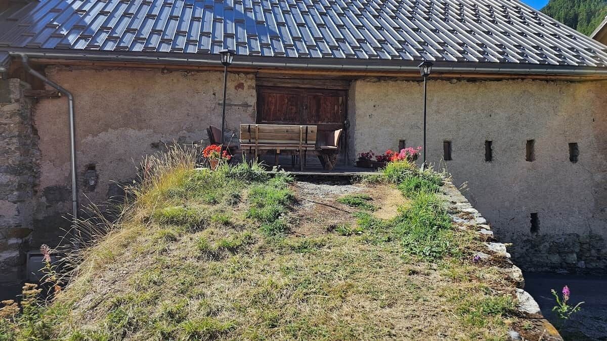 Un edificio rústico con techo de tejas, una banca de madera en el exterior y un pequeño jardín.