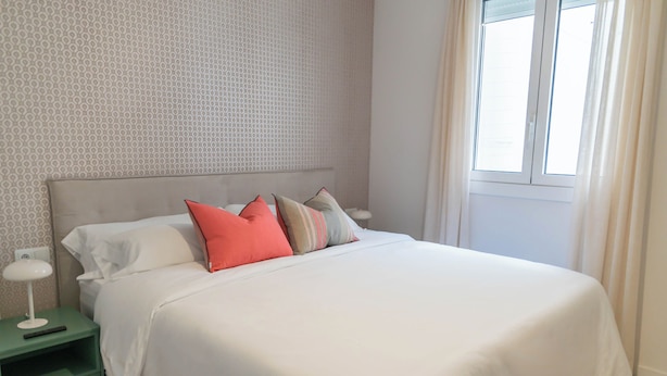 Una cama bien hecha con ropa blanca y varios almohadones, una mesita de noche verde con una lámpara, y una ventana con cortinas.