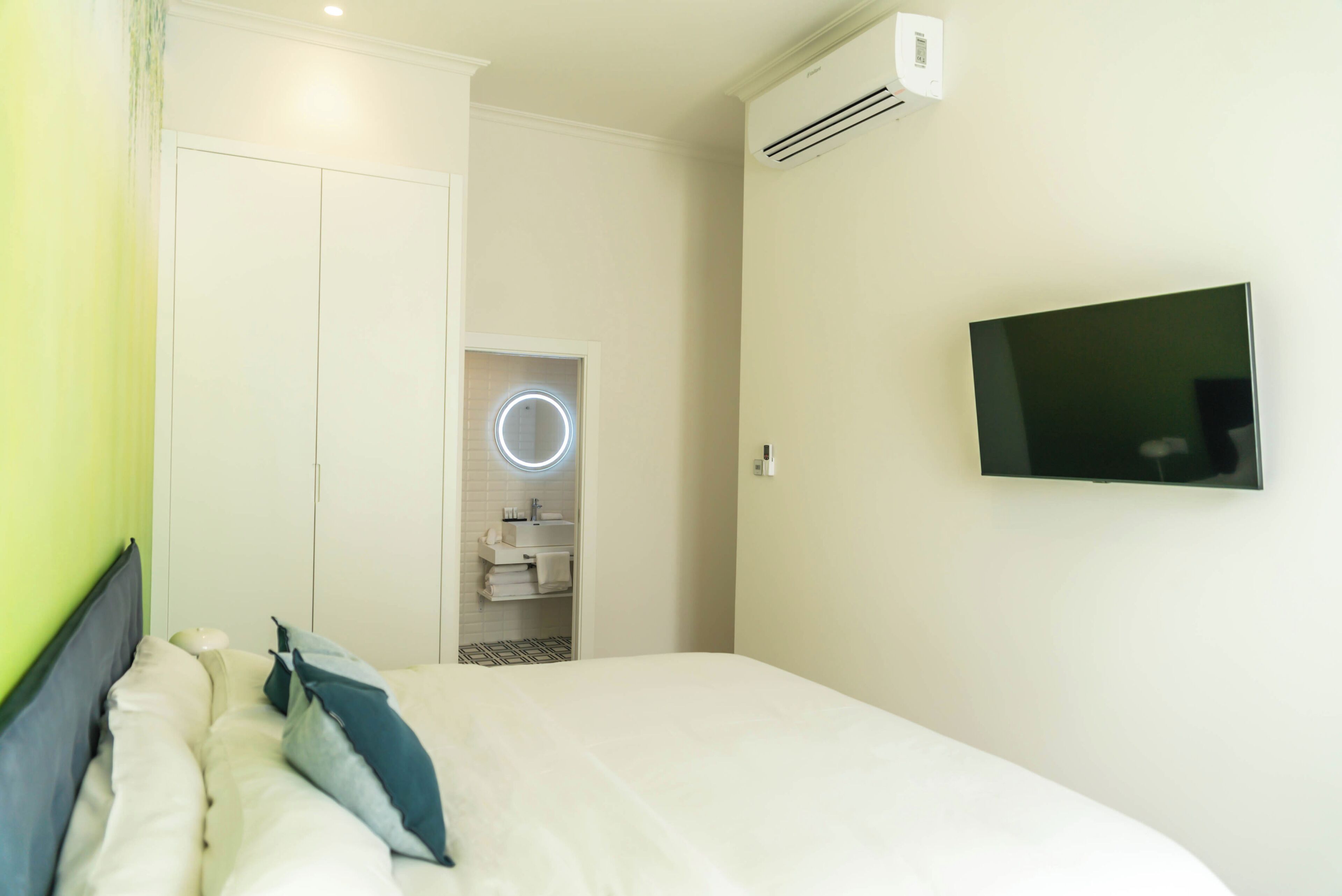 Et hotelværelse med en seng, et fjernsyn ophængt på væggen, et badeværelse med et rundt spejl og en aircondition.