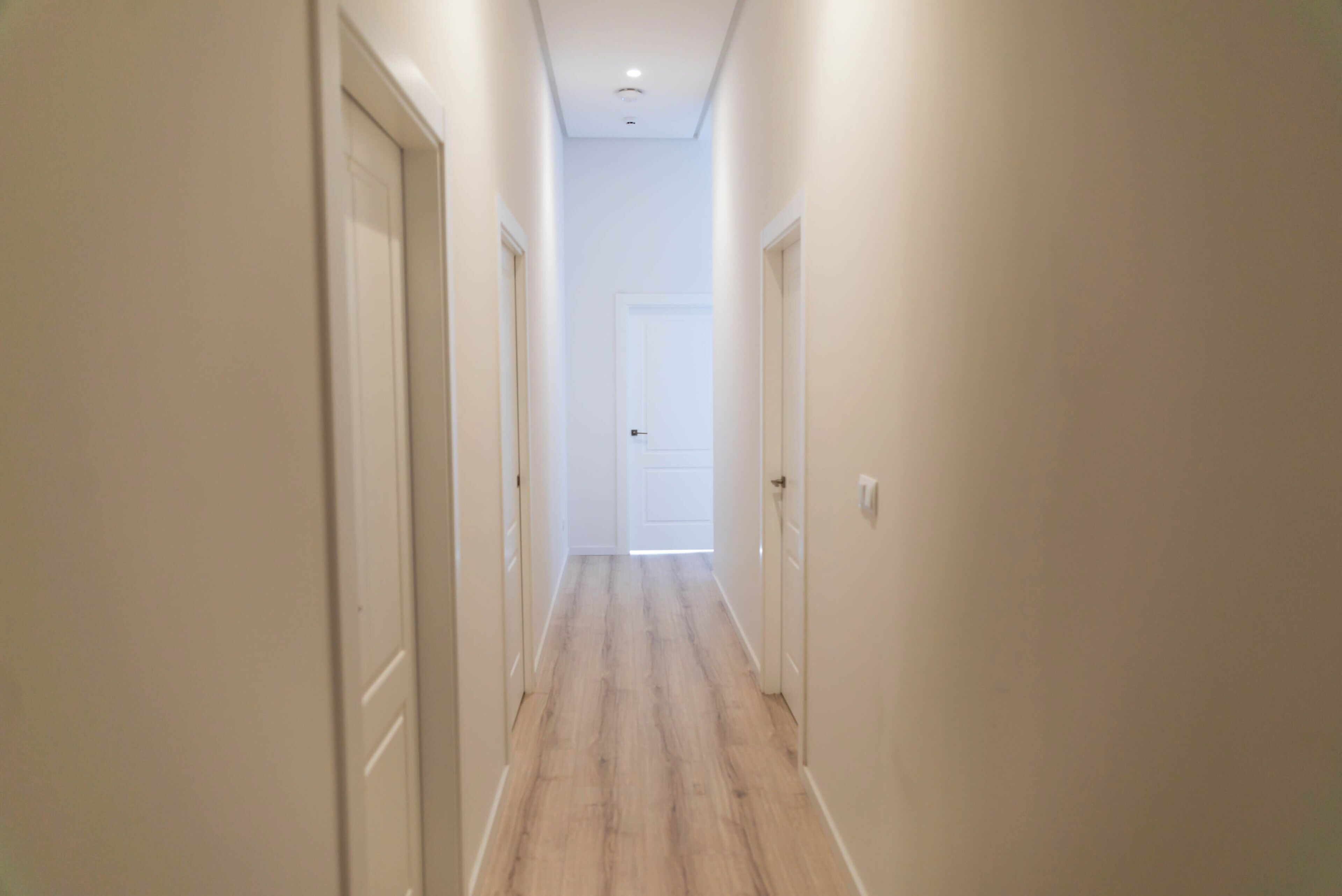 A narrow hallway with wooden flooring and white walls, leading to a closed door at the end.