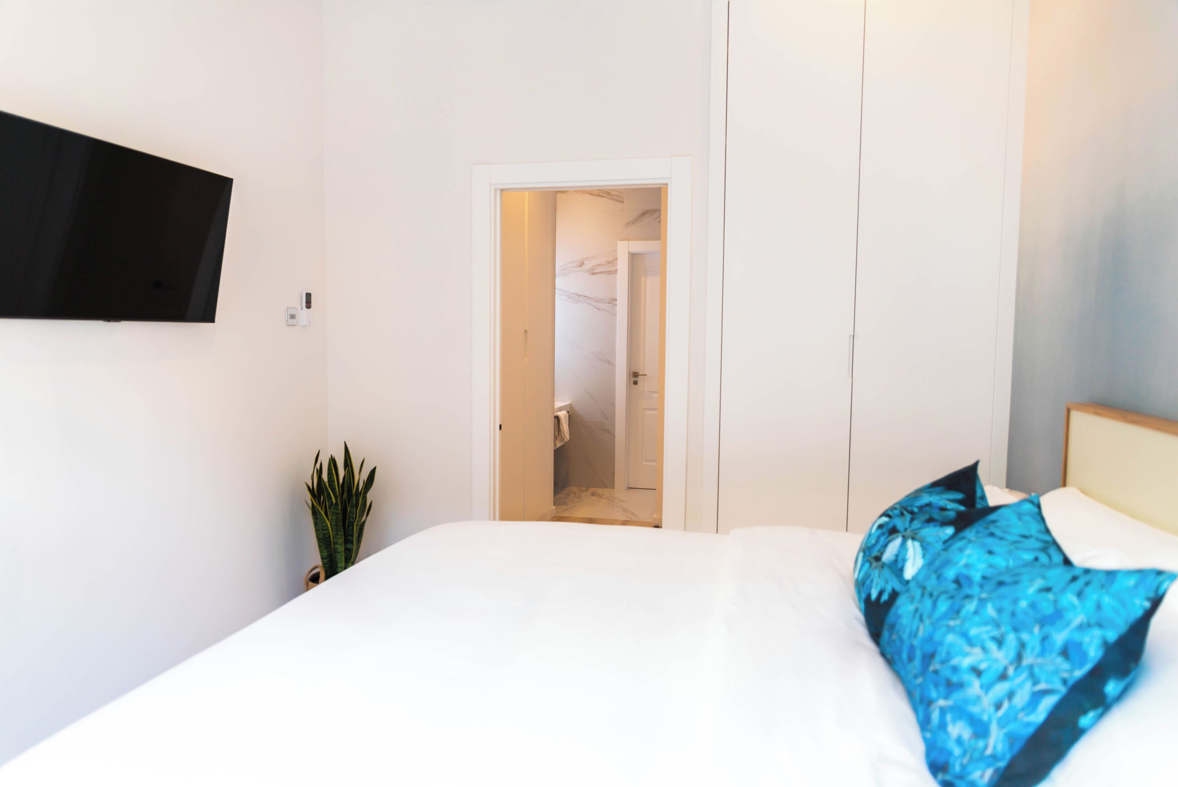 A bedroom with a bed, a TV mounted on the wall, a potted plant, and a door leading to a bathroom.