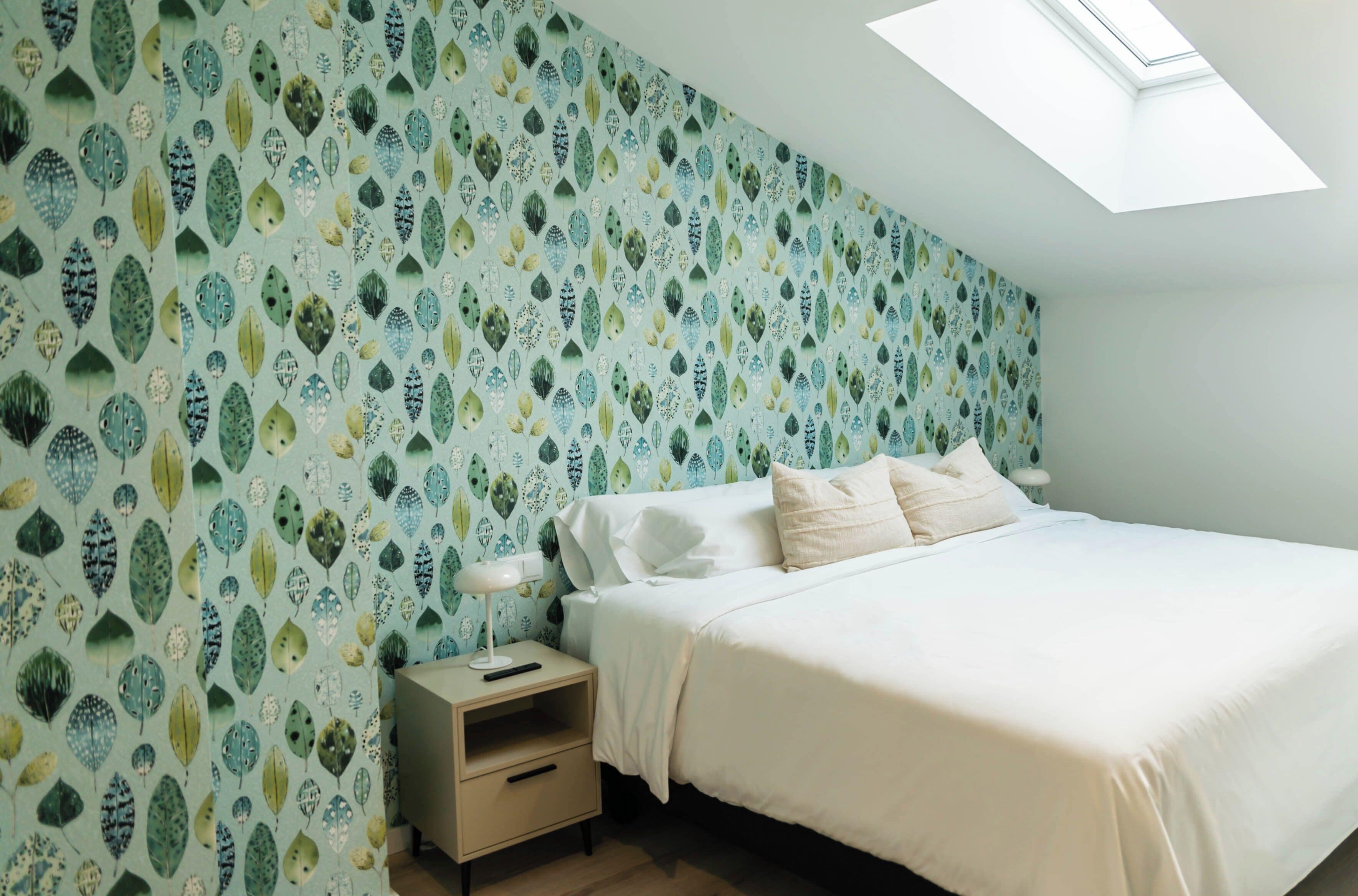 Un dormitorio con techo inclinado, tragaluces, cama con ropa de cama blanca, mesita de noche con lámpara y pared con diseño de hojas.