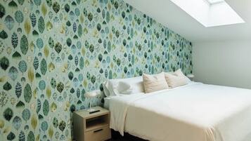 Un dormitorio con techo inclinado, tragaluces, cama con ropa de cama blanca, mesita de noche con lámpara y pared con diseño de hojas.