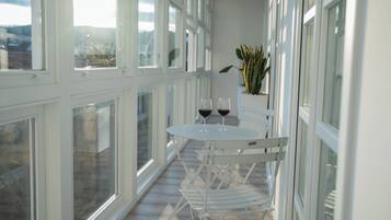 Un balcón con una mesa blanca y dos copas de vino tinto, rodeado de sillas blancas.