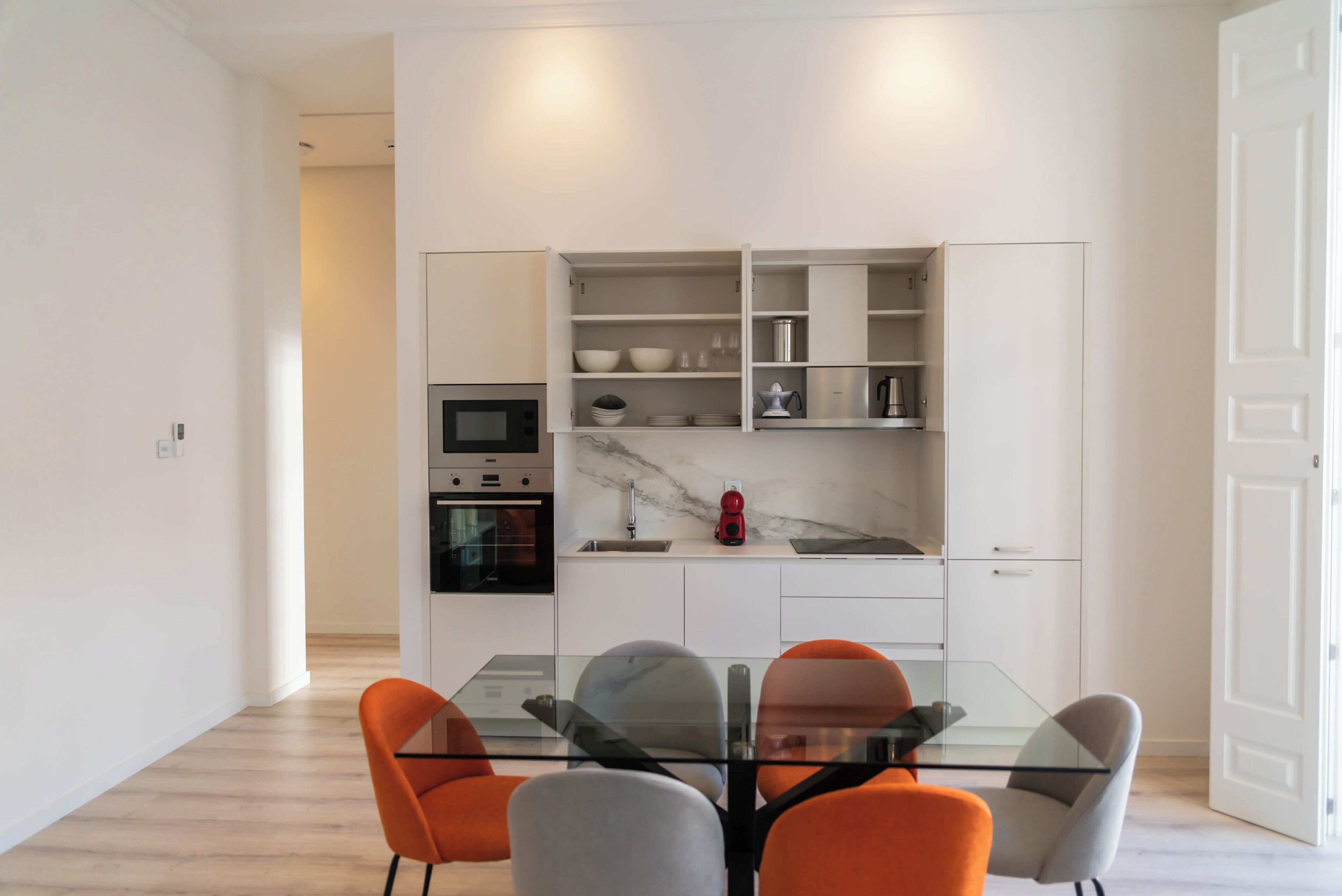 Une cuisine moderne avec des meubles blancs, un four encastré et une table à manger en verre, assortie de chaises orange.