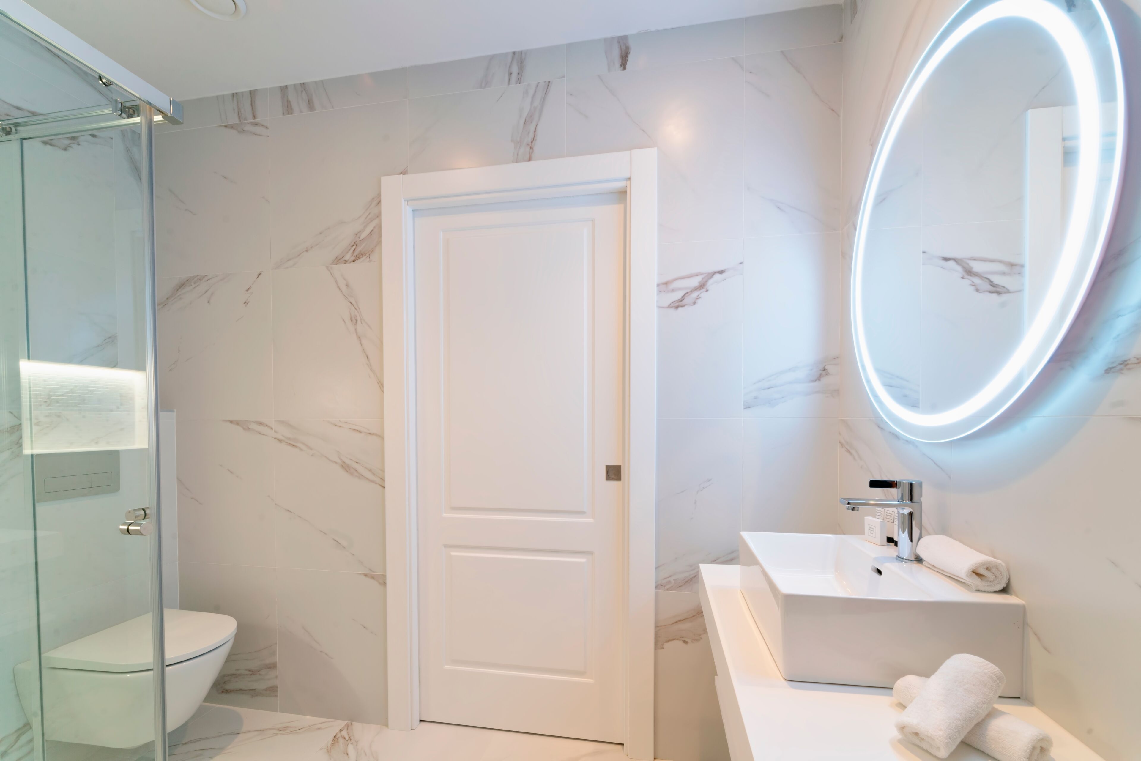 A modern bathroom with a glass shower enclosure, a white toilet, a marble-tiled wall, and a vanity with a mirror.