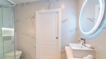 A modern bathroom with a glass shower enclosure, a white toilet, a marble-tiled wall, and a vanity with a mirror.