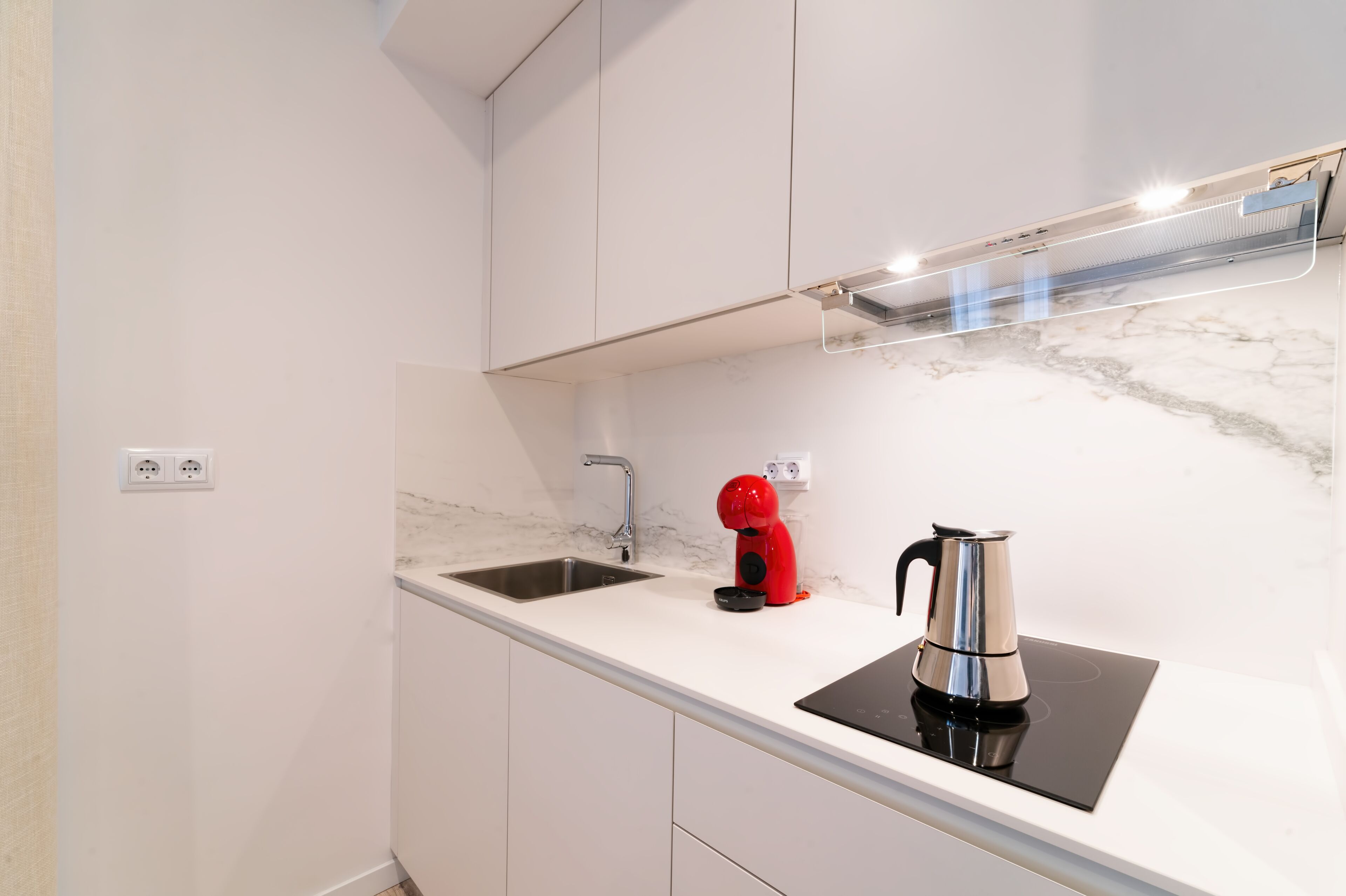 Cocina moderna con encimera blanca, pileta de acero inoxidable, cafetera roja y cocina por inducción negra.