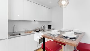 A modern kitchen with white cabinets, a stainless steel sink, a microwave, and a dining area with a wooden table and red chairs. - Morar Apartments Malasaña (Madrid)