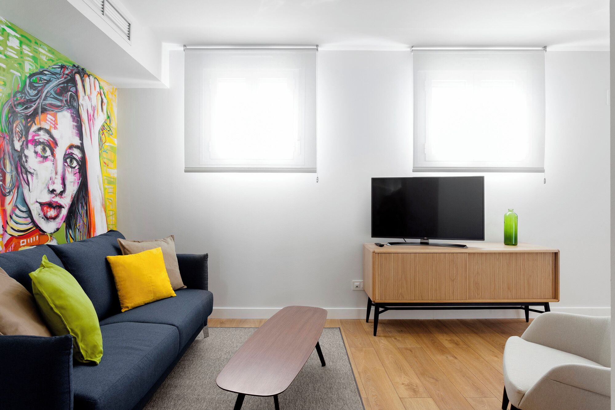 Ein modernes Wohnzimmer mit einer Couch, einem Couchtisch, einem Fernseher auf einer Holzkommode und einem farbenfrohen Gemälde an der Wand.