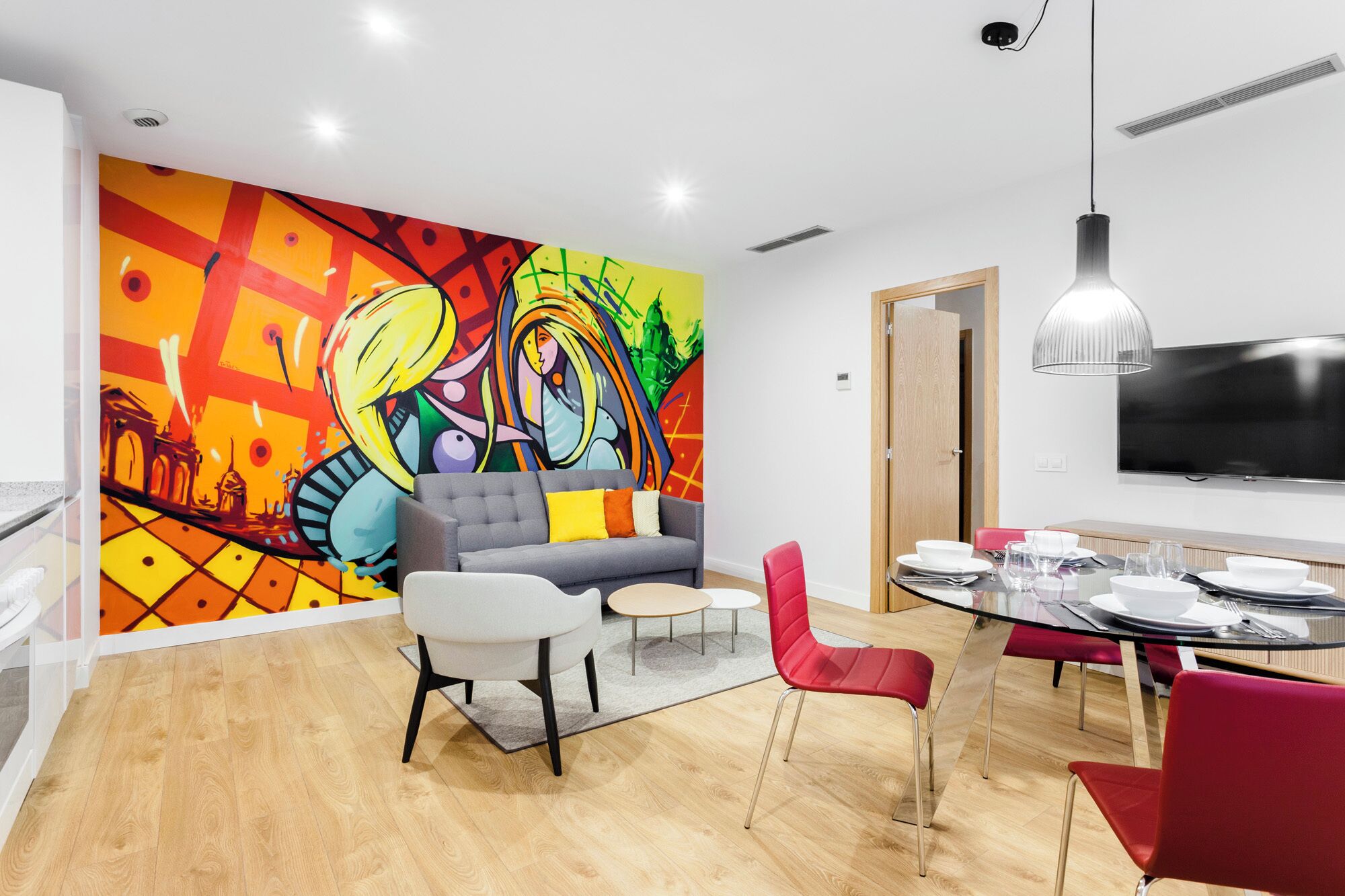 A modern living room with a colorful mural, a grey sofa, red chairs, a dining table with chairs, and a flat-screen TV.