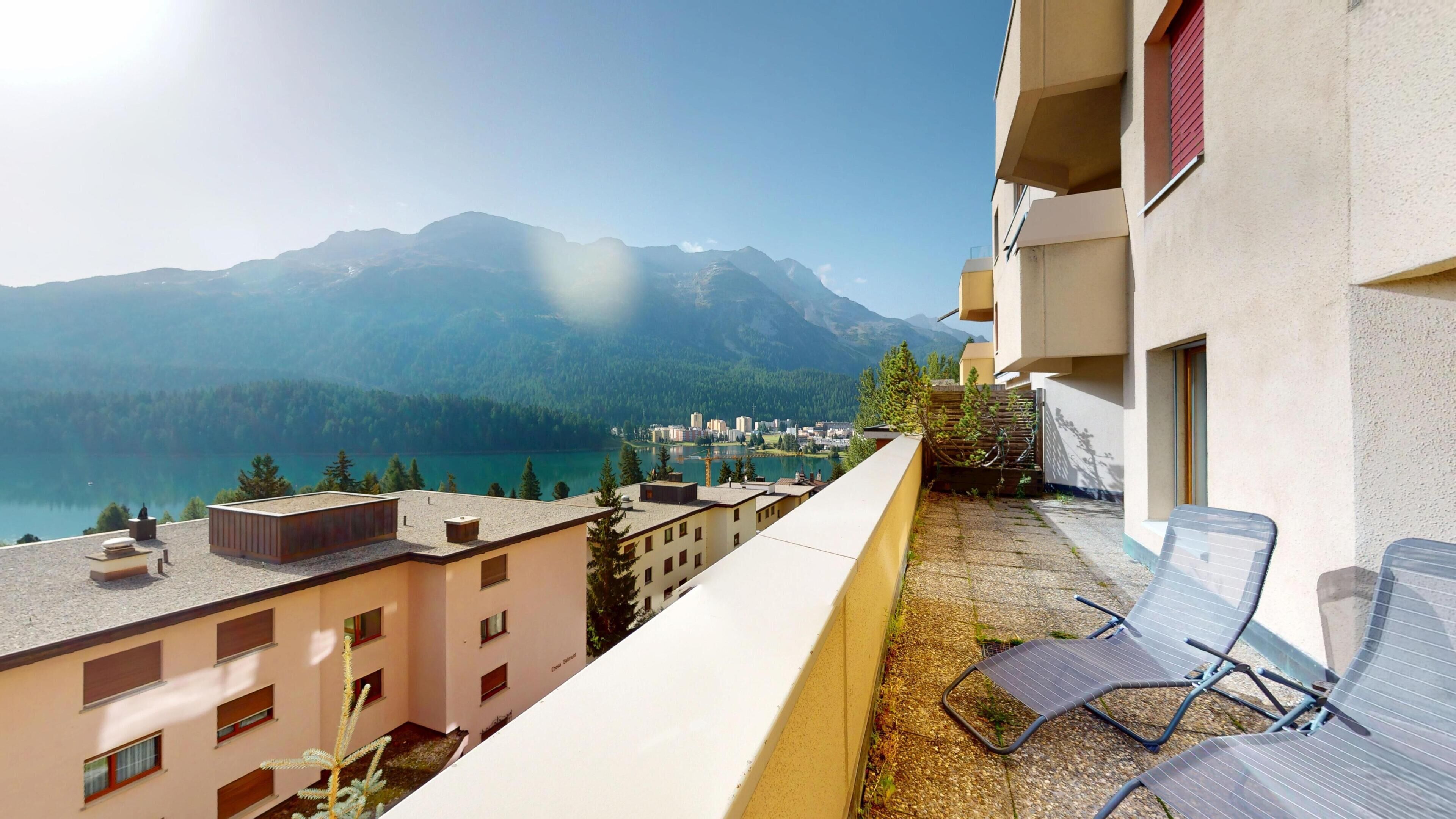 A balcony with a view of a lake and mountains, a chair, and a building with a balcony.