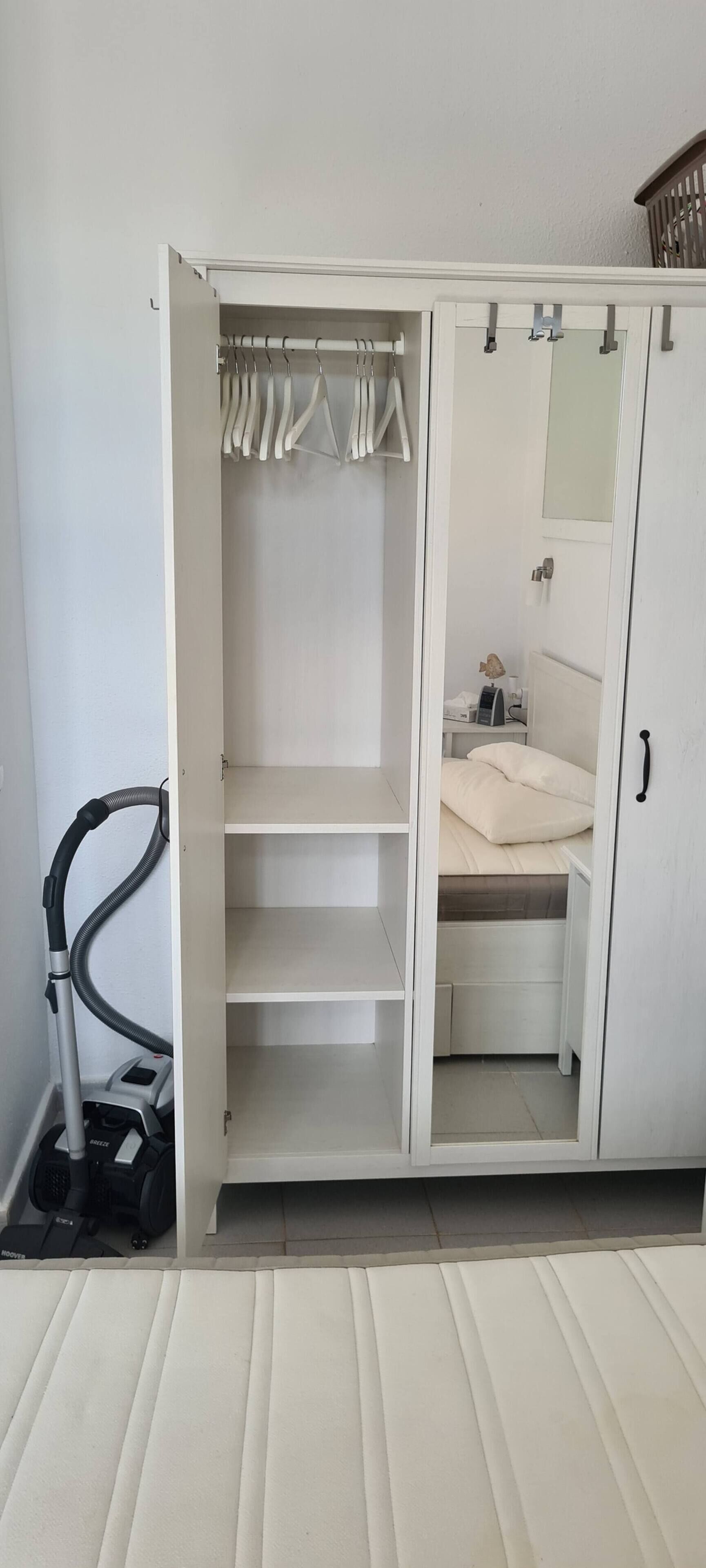 Ein weißer Kleiderschrank mit Spiegel, ein Bett mit weißer Matratze und ein Staubsauger.