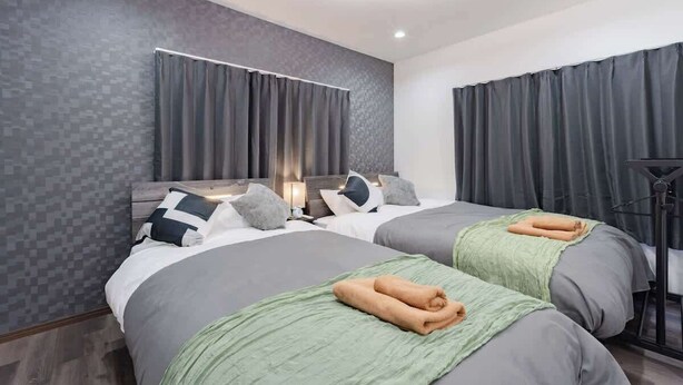 Une chambre d’hôtel avec deux lits, une literie grise, un plaid vert, une table de chevet avec une lampe et un luminaire moderne au plafond.